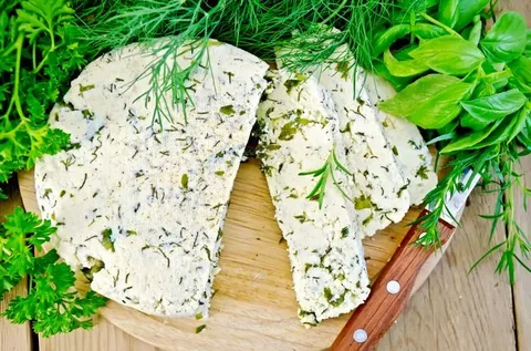 “Doğal otlarla hazırlanmış taze otlu peynir dilimleri, yeşillikler ve bıçakla servis tahtası üzerinde.”